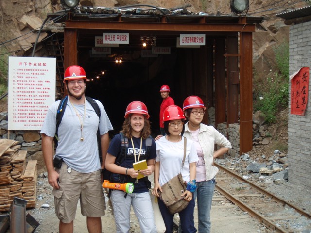 Four students standing in front of the entrance tunnel of
          a Chinese molybdenite mine in Henan Province. The students
          have red miner's hard hats on their heads and big flashlights
          to see in the dark. Signs posted on the sides of the tunnel
          entrance are written in red Chinese characters.
